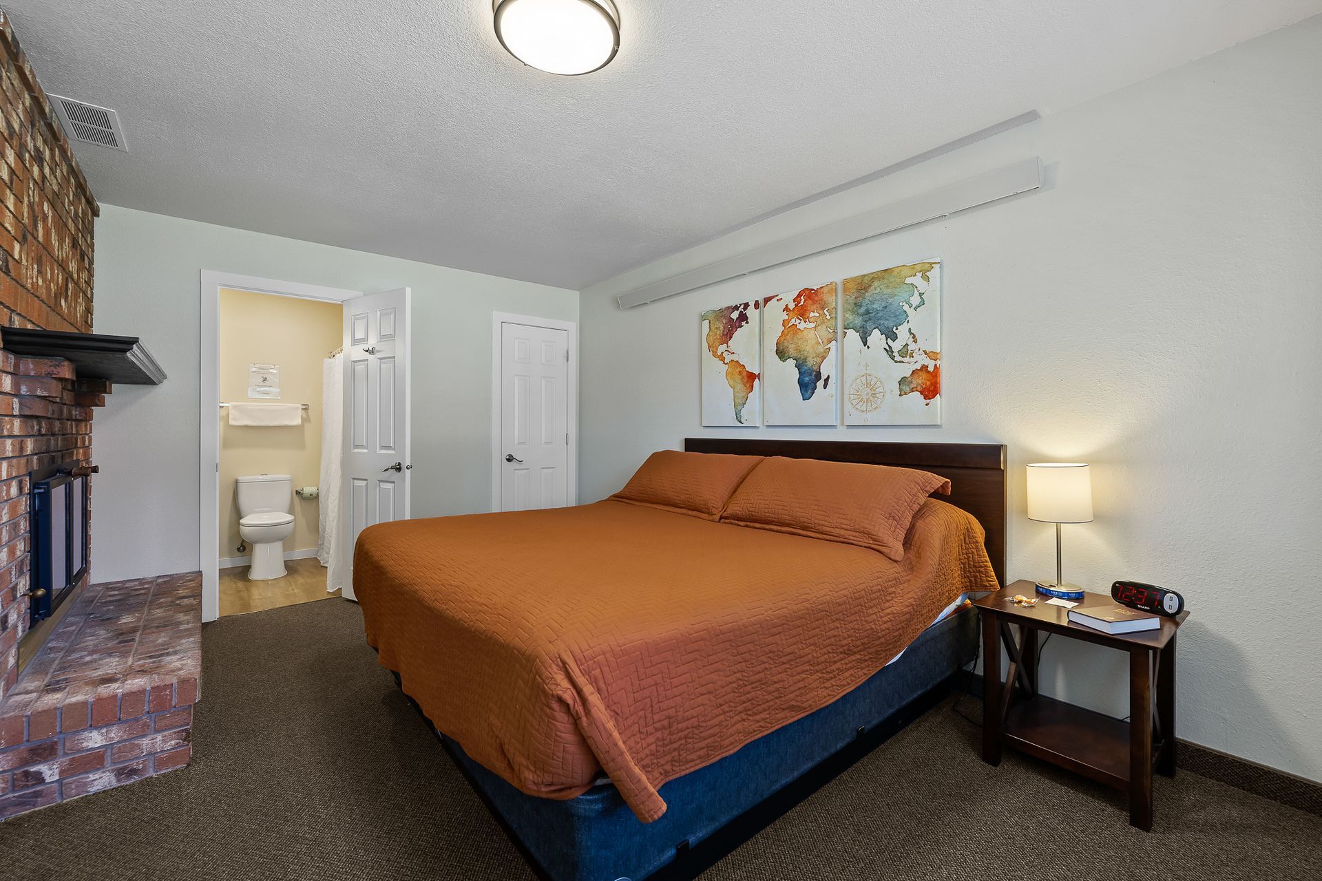 Bedroom with queen bed, fireplace, and world map artwork. Small nightstand with lamp. Toilet visible in open doorway.