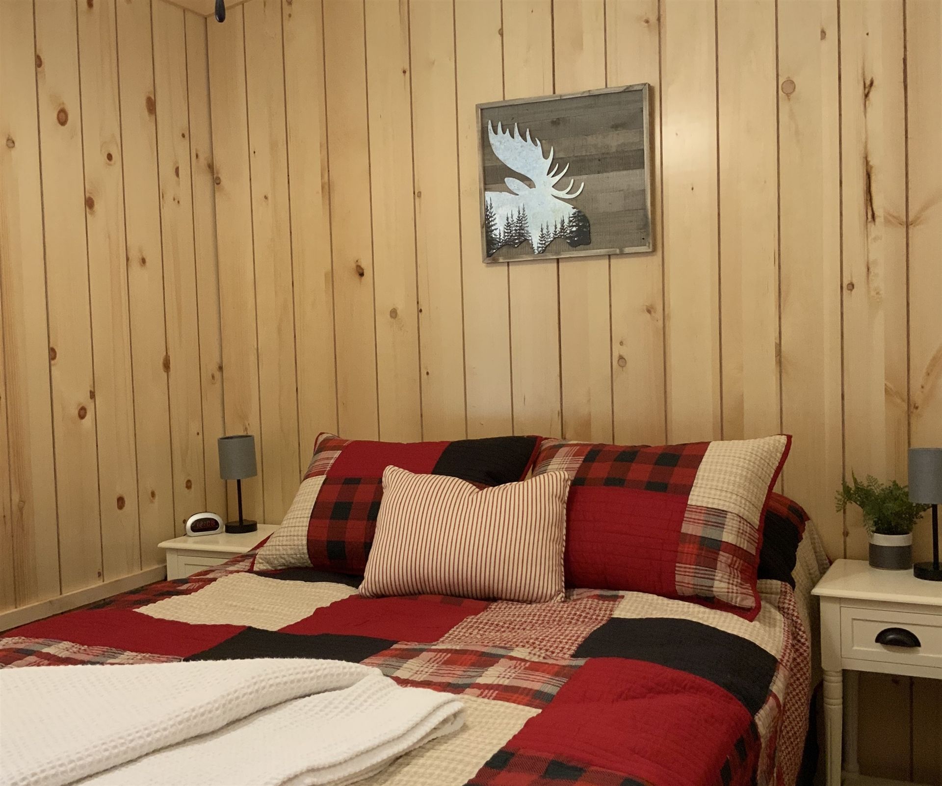 Bedroom with red, black, and cream patchwork bedding, wood-paneled walls, and a moose art piece above the bed.