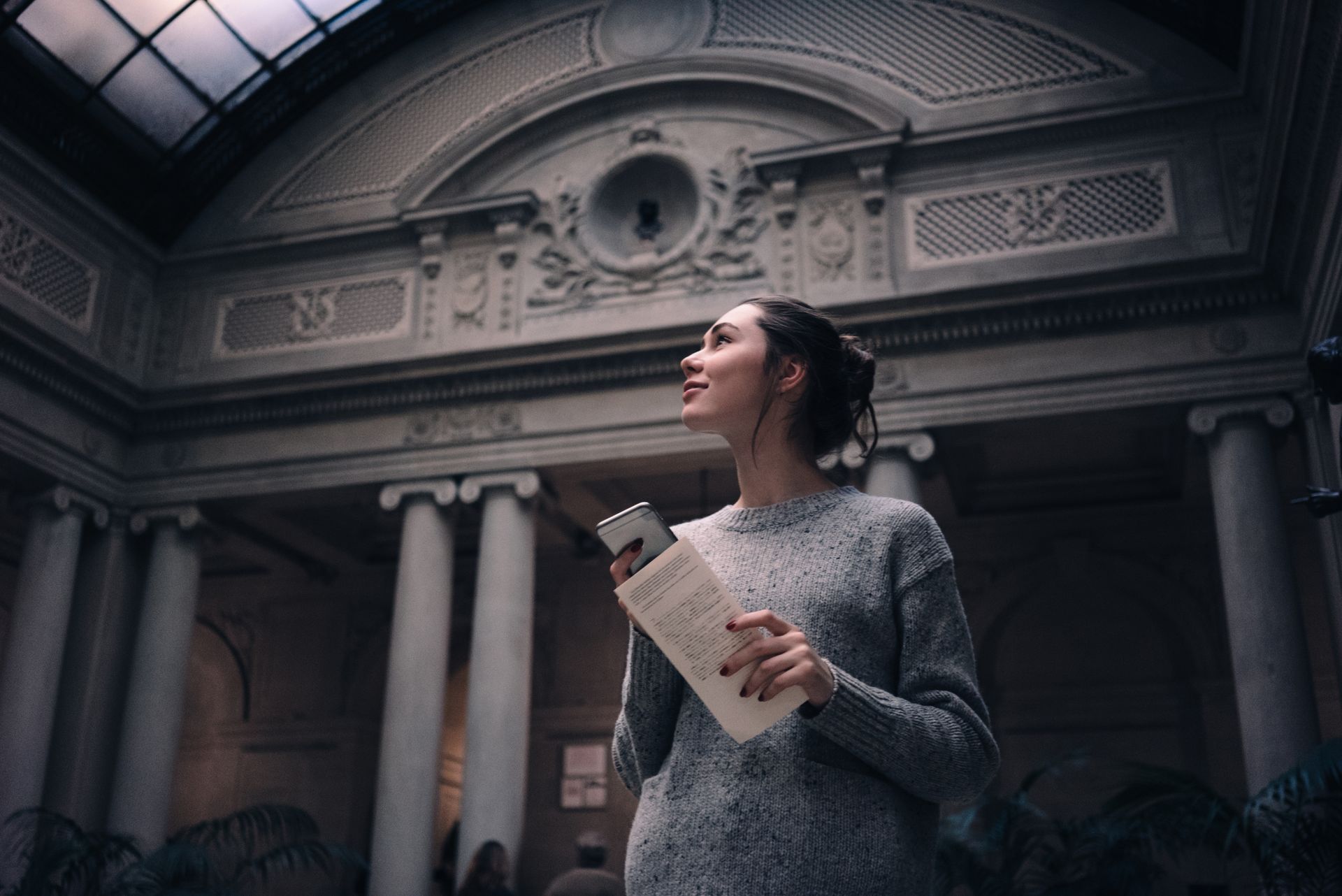 Woman holding a paper in a grand stone hall, looking upward beneath ornate arches.