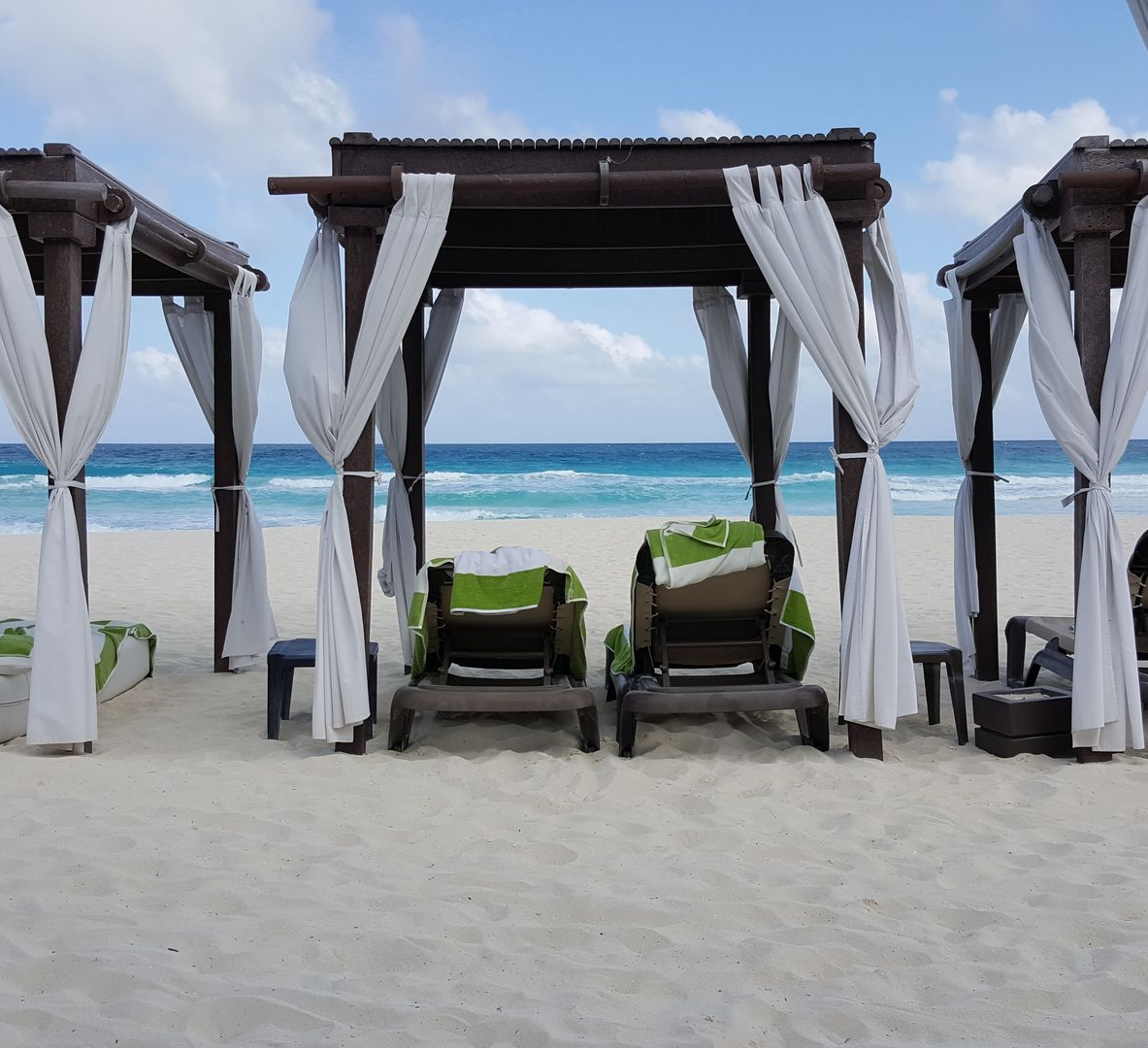 Beach cabanas with white drapes and lounge chairs facing a turquoise ocean under a partly cloudy sky
