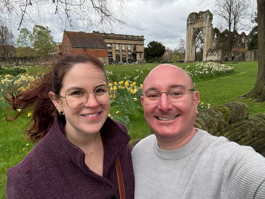 Two smiling people taking a selfie in a grassy park with old stone ruins and a building behind them.