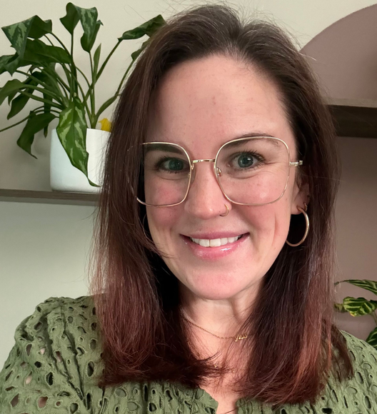 Smiling woman with glasses and hoop earrings in a green top, standing indoors near plants.