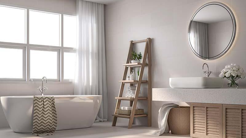 Bright bathroom with freestanding tub, ladder shelf, round mirror, and white vanity by a window