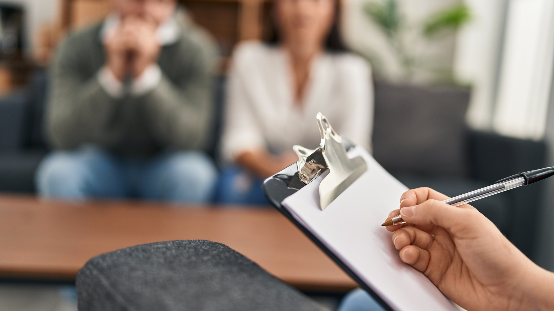 Two people sit on a couch, holding hands in a counseling session near a bright window.