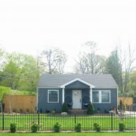 Small blue-gray house with a front porch, black fence, and young shrubs in a yard.