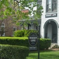 Biscuit Love restaurant storefront with arched doorway, hedges, and hanging sign on a tree-lined street
