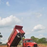Red dump truck tipping its bed to unload soil at a construction site under a blue sky.