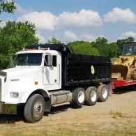 White dump truck with black bed hauling heavy equipment on a trailer in a dirt lot under a blue sky
