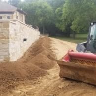 Construction equipment grading dirt beside a stone building and trees