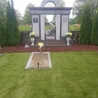 Memorial garden with headstone monument, flowers, and manicured lawn under overcast sky
