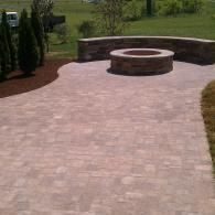 Stone-paved patio with a circular fire pit and curved stone seating wall overlooking a green lawn