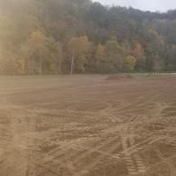 Recently plowed field with tire tracks, bordered by autumn trees and a wooded hillside.