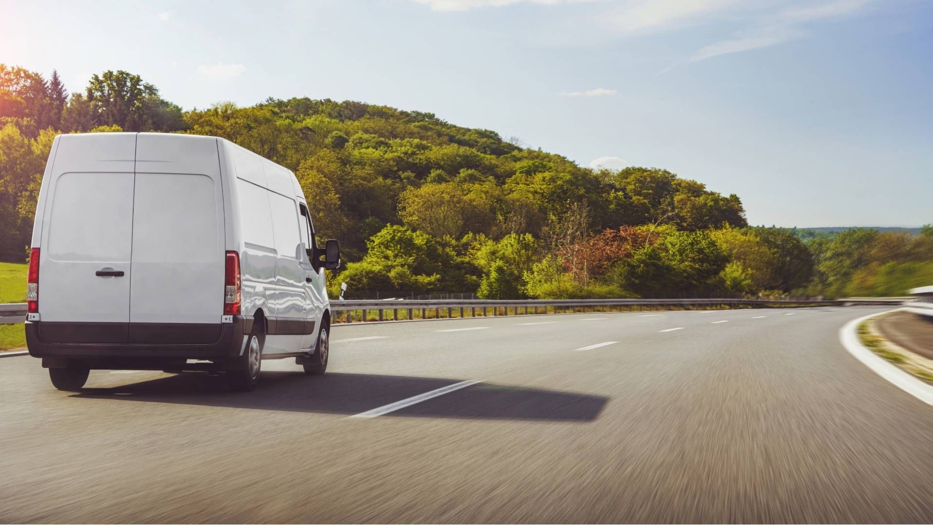 White cargo van driving on a curving highway beside green hills on a sunny day
