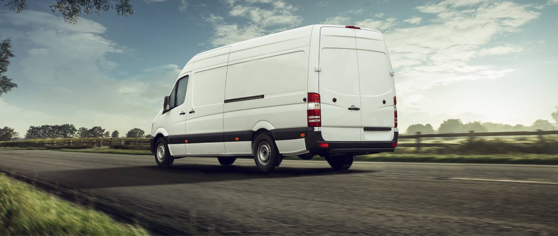 White cargo van driving on a road beside grassy fields under a cloudy sky
