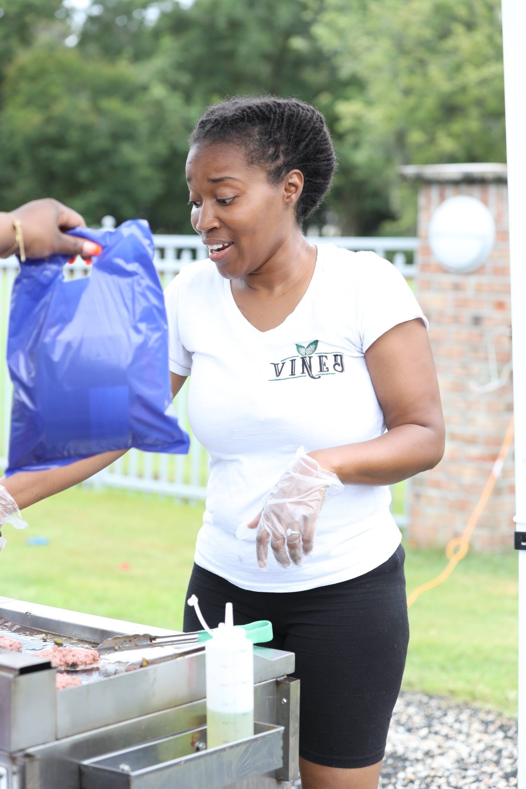 Woman in white shirt handing a blue bag at an outdoor food-serving table.