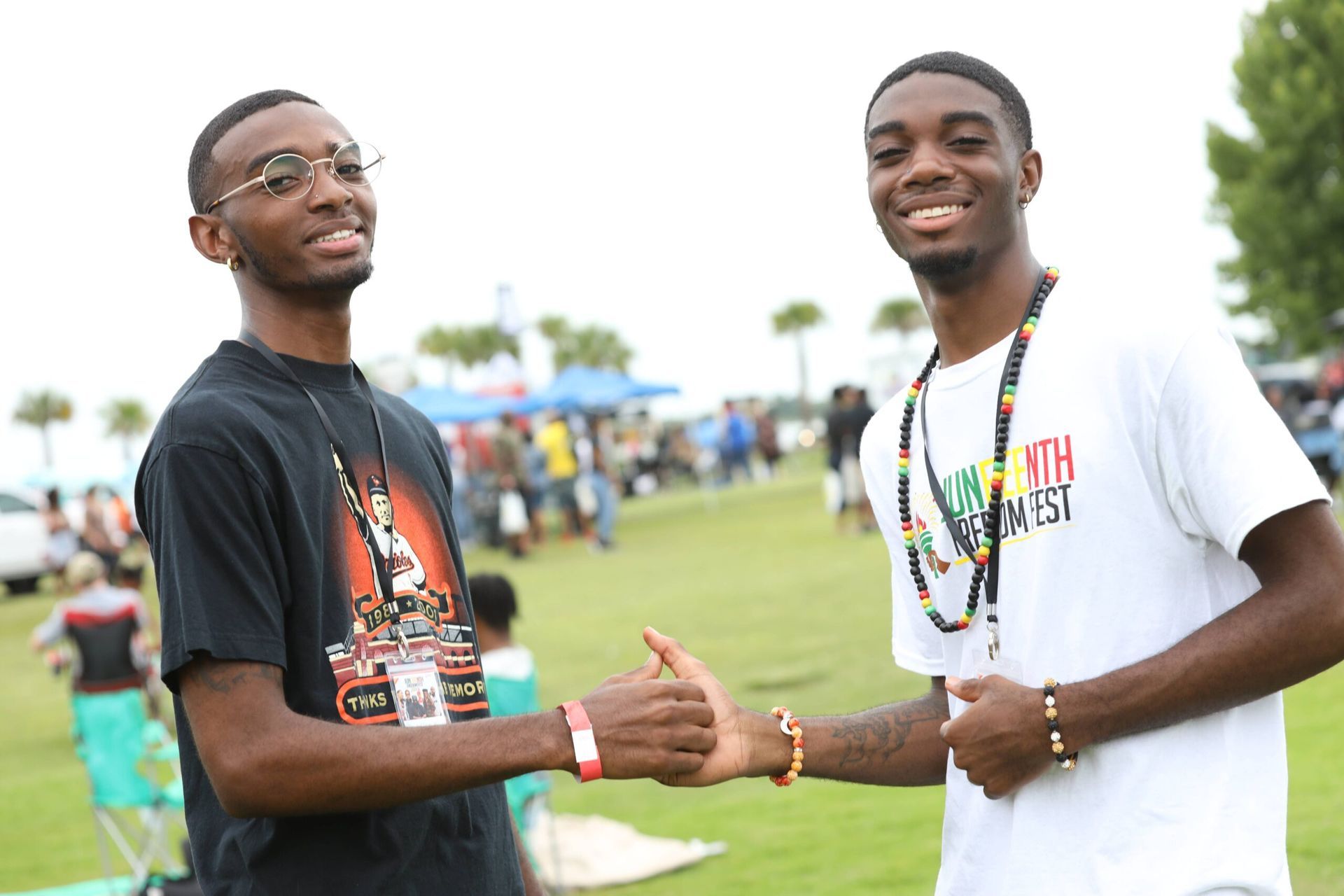 Two men smiling and shaking hands outdoors at an event, wearing lanyards and casual T-shirts