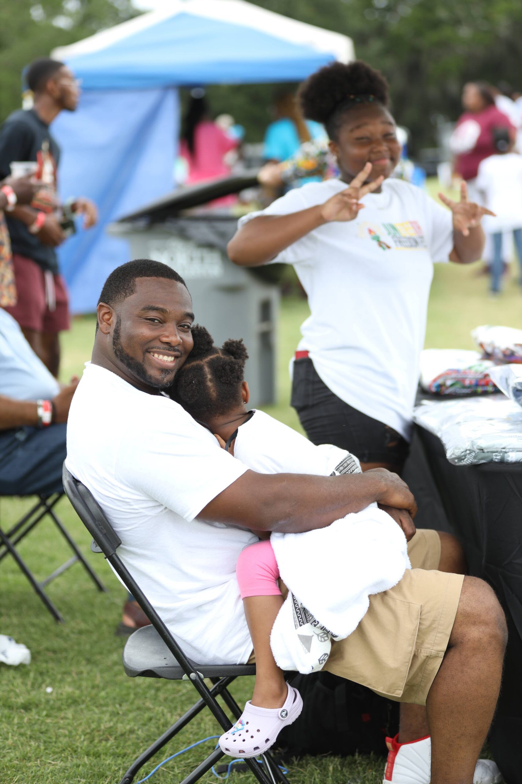 Smiling man in white shirt sitting outdoors, hugging a child, with another child standing nearby at a picnic table