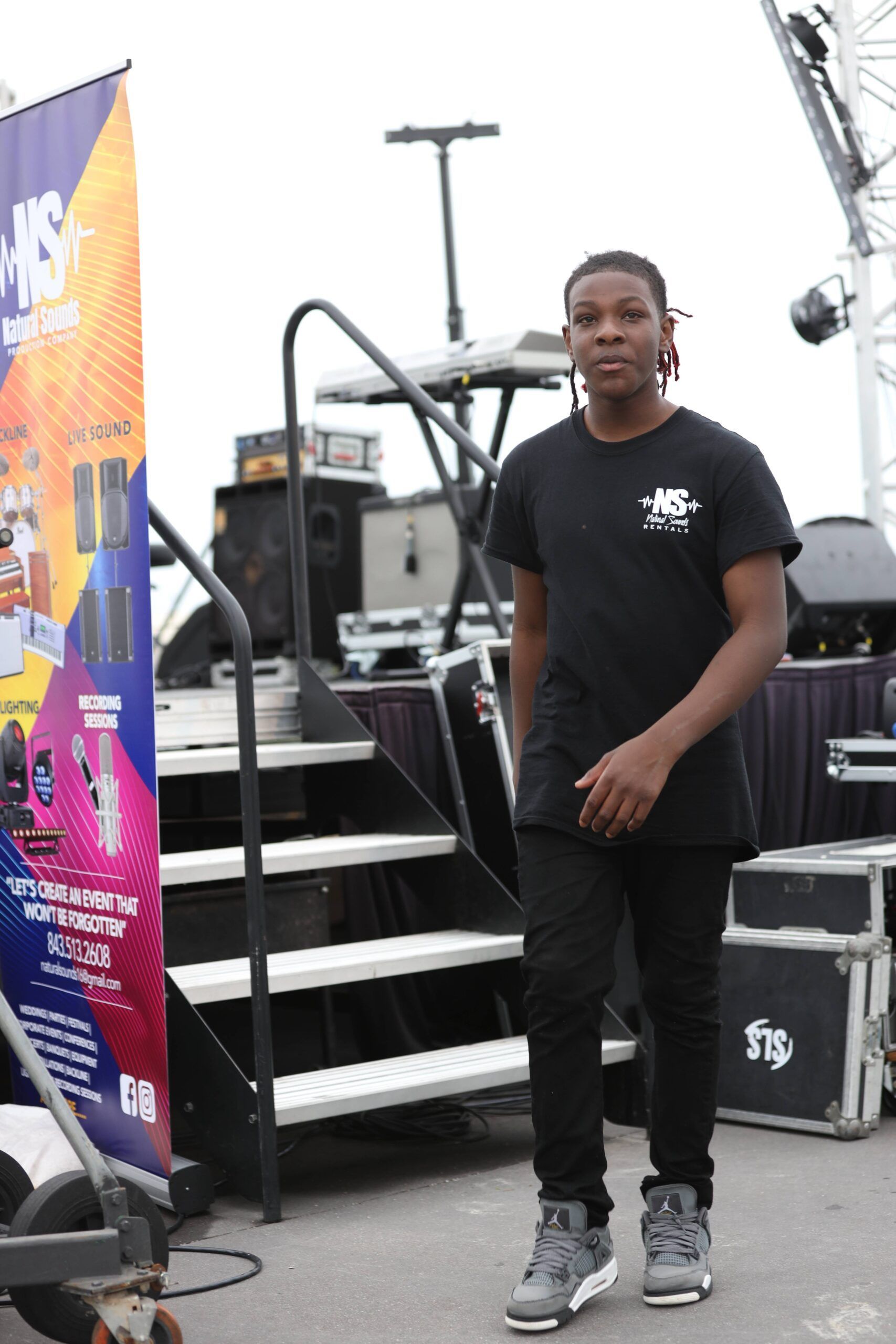 Person standing on outdoor stage near stairs and equipment, wearing a black T-shirt and gray sneakers.