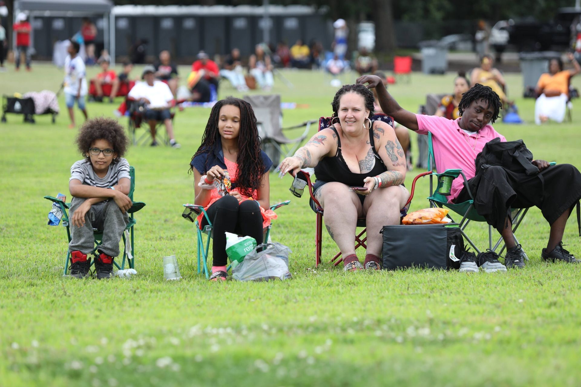 People sitting on folding chairs in a grassy park, relaxing near a crowd in the background.