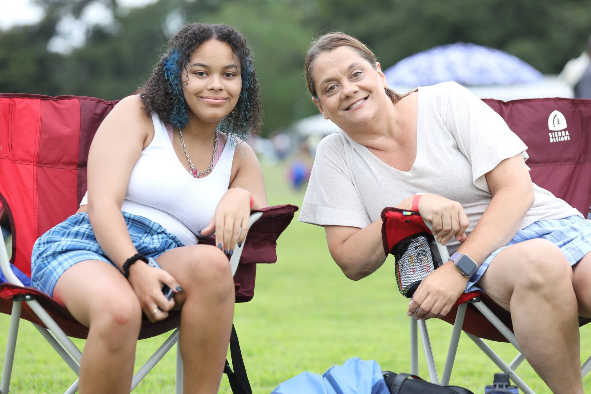 Two people smiling in camping chairs on a grassy field with tents behind them.