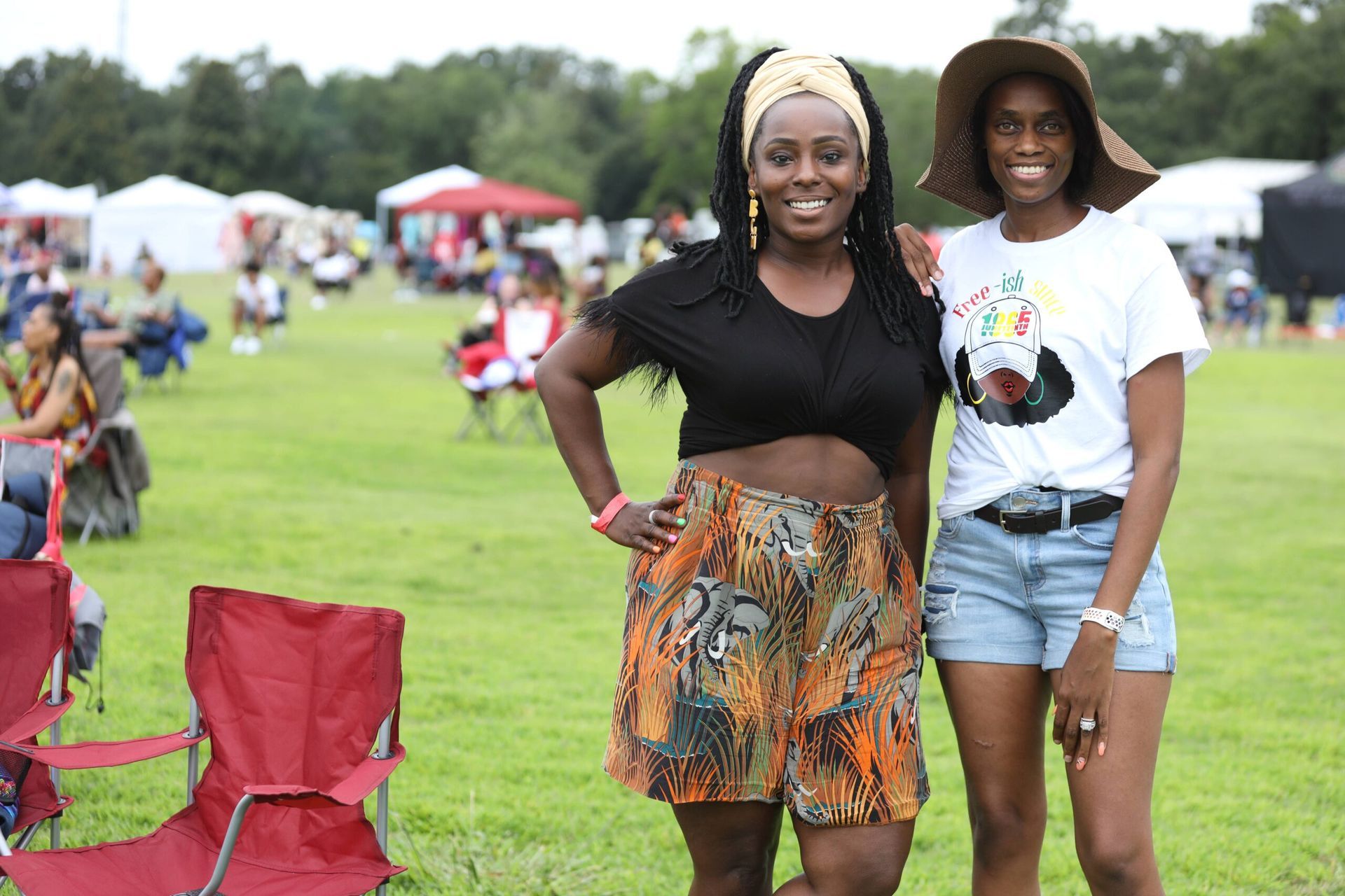 Two women smiling at an outdoor festival, standing on grass near red chairs and tents.