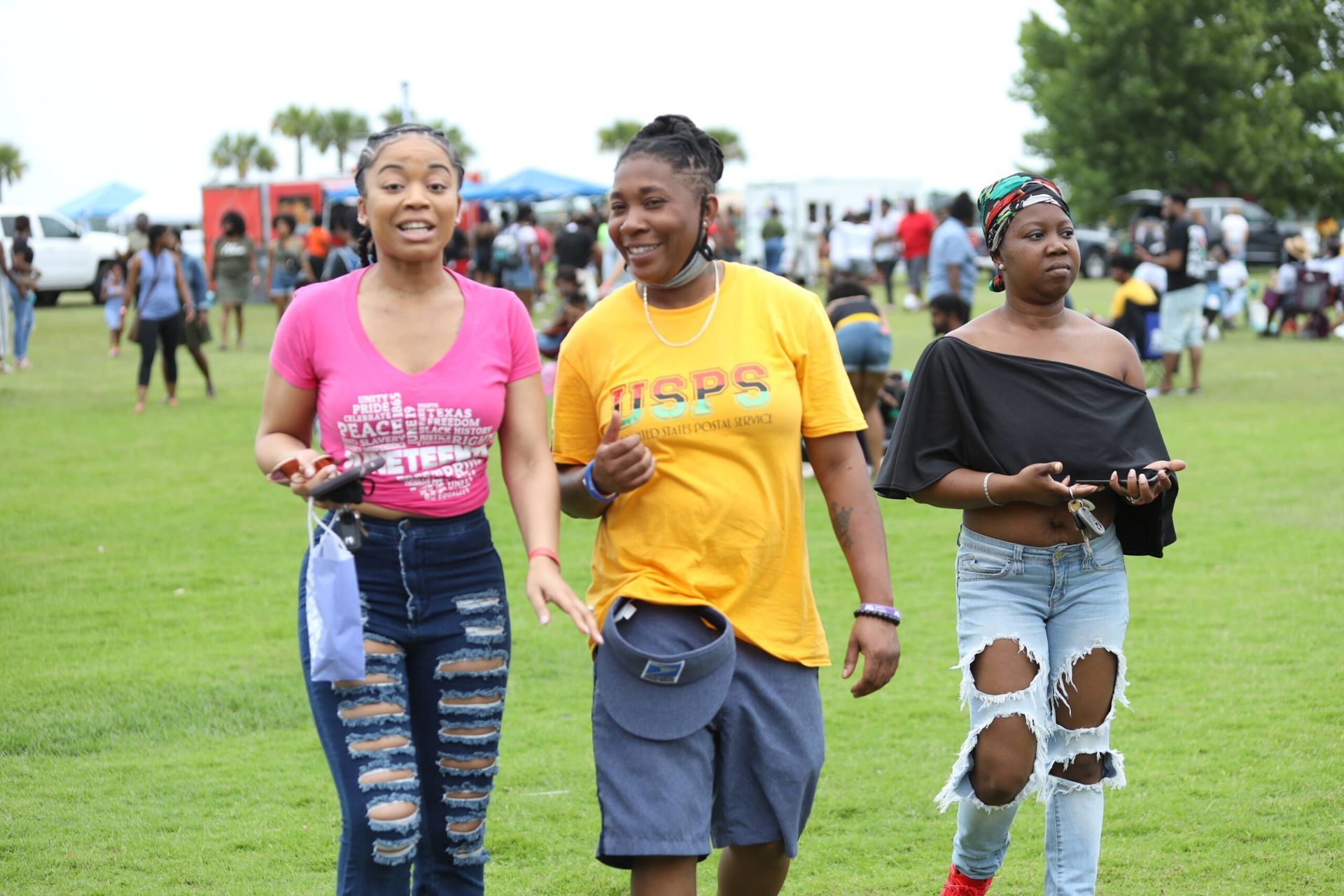 Three people walking on a grassy field, smiling, with a crowd in the background.