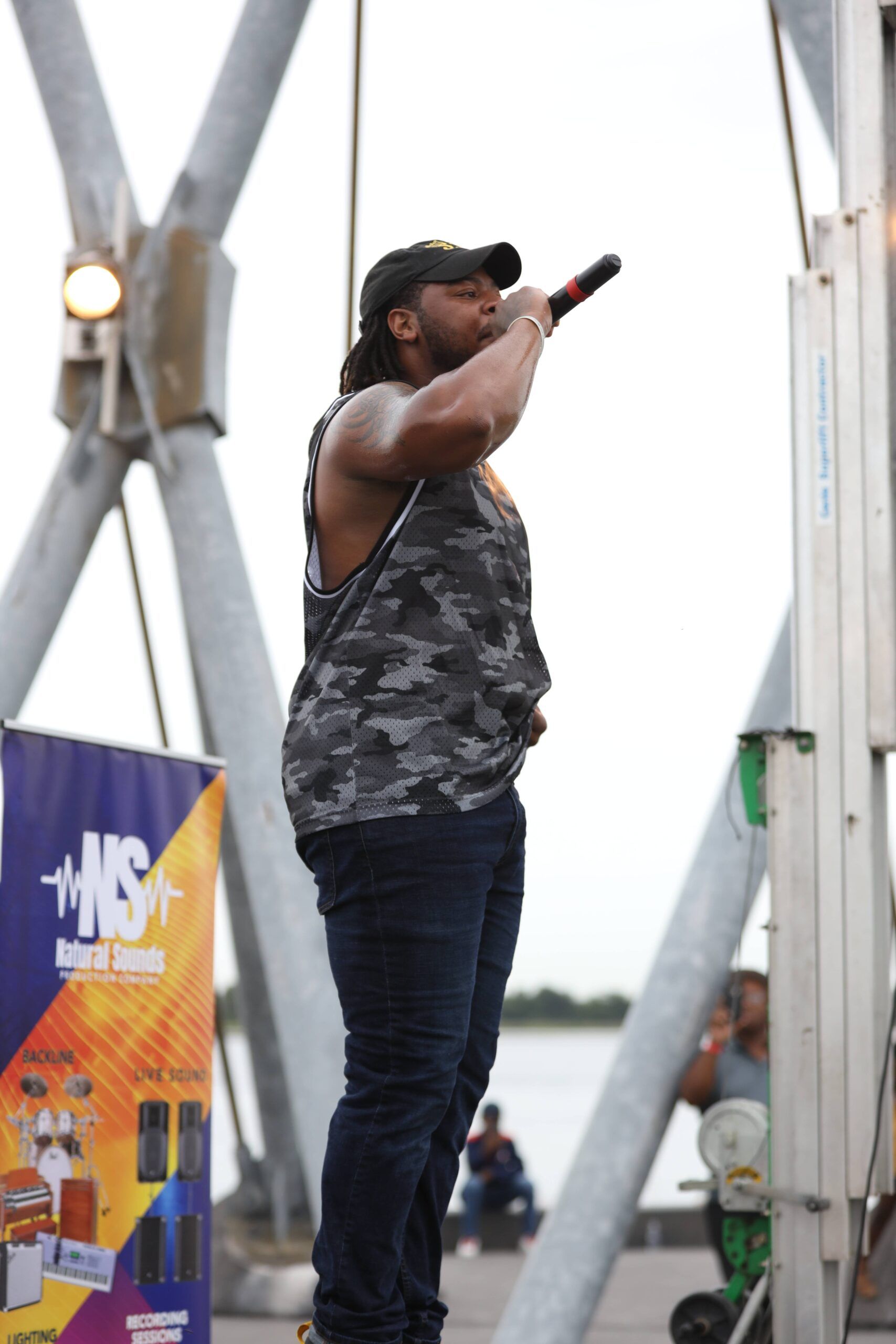 Singer performing on an outdoor stage with a microphone, wearing a black cap and camo tank top.