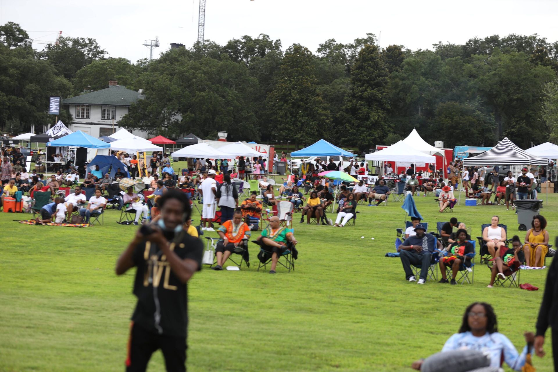 Crowd seated on a grassy field at an outdoor festival with tents and a photographer in the foreground
