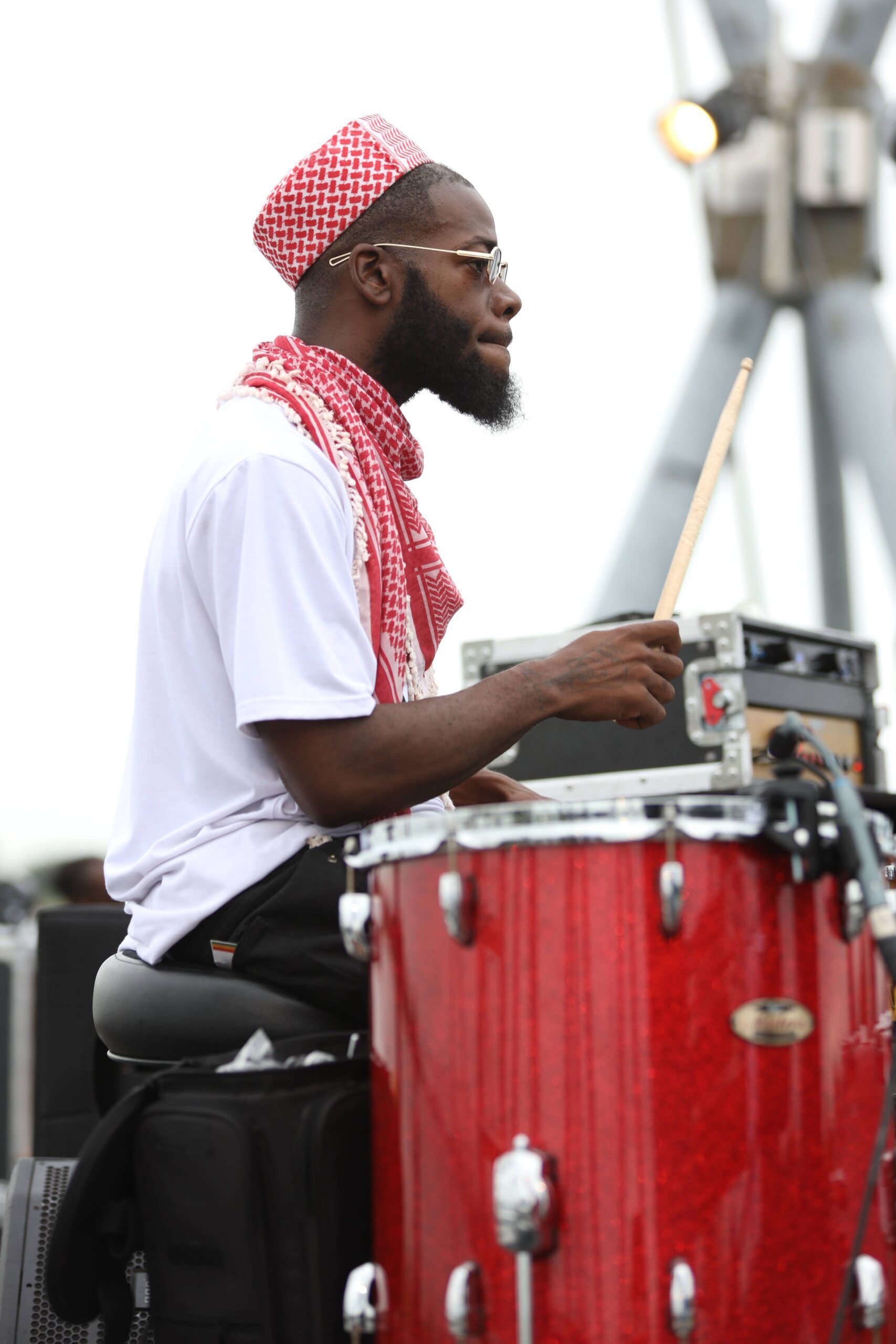 Drummer playing red drums on an outdoor stage, wearing a white shirt and red patterned headscarf.