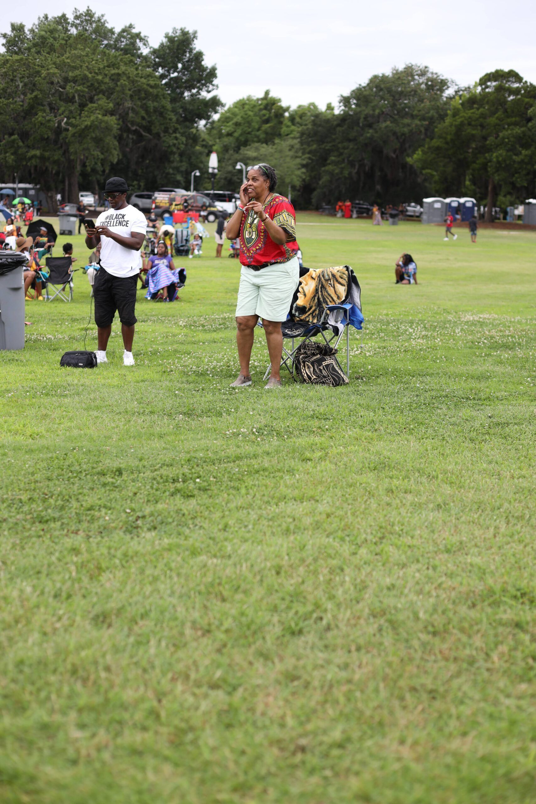 People standing on a grassy field at an outdoor park event, with trees and tents in the background.