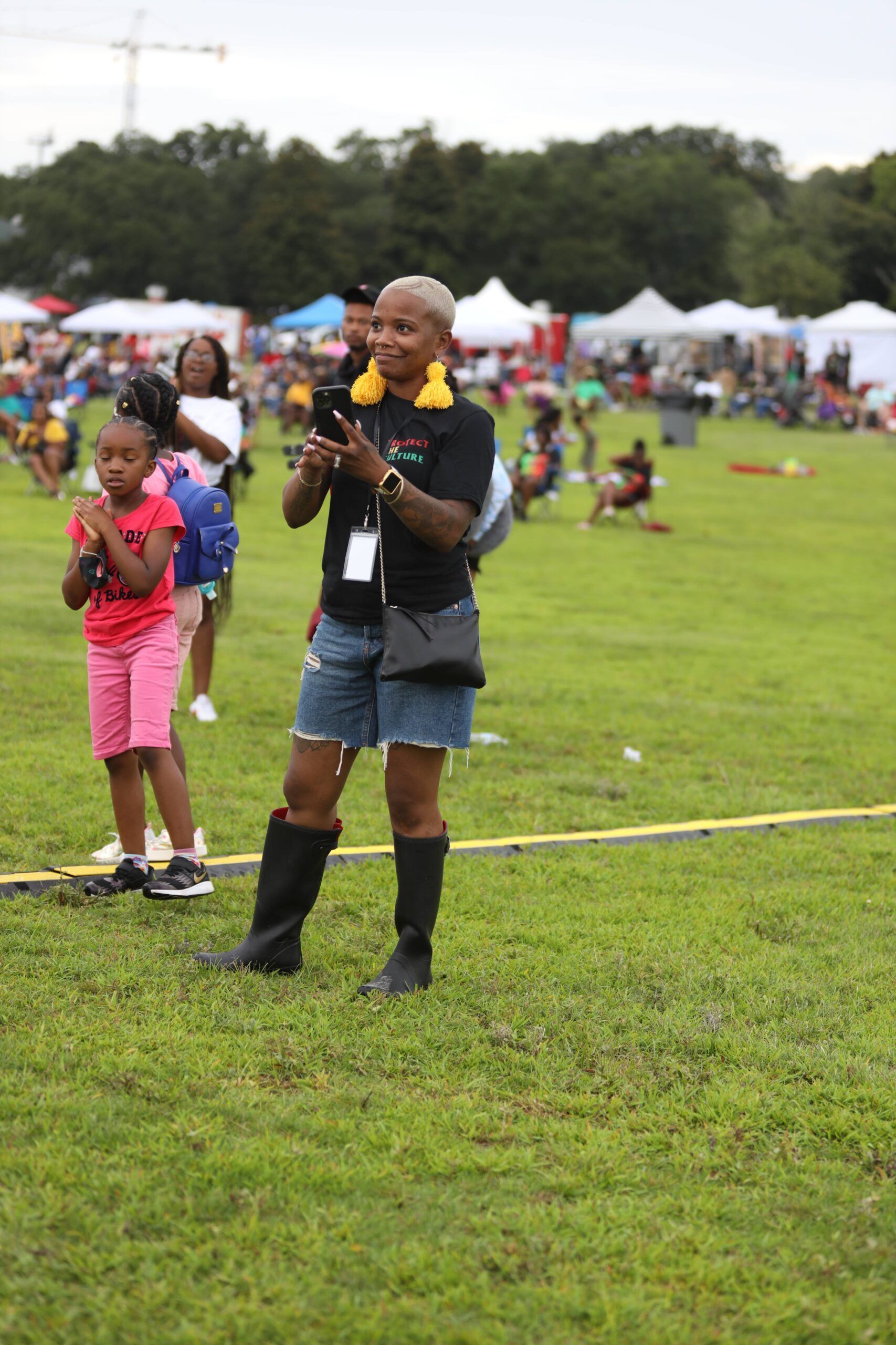 Person holding a baby with a child nearby at an outdoor grassy event with tents in the background