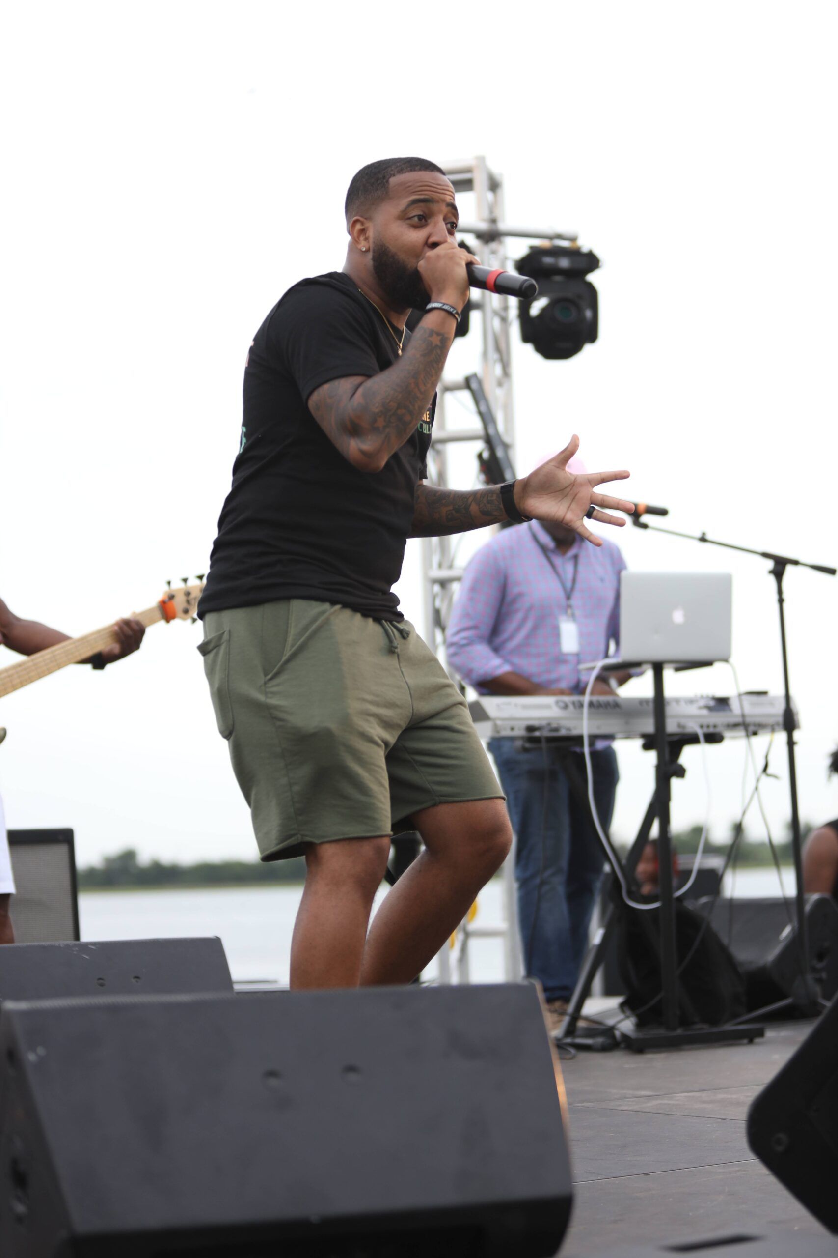 Singer performing on an outdoor stage with a microphone, wearing a black shirt and green shorts.