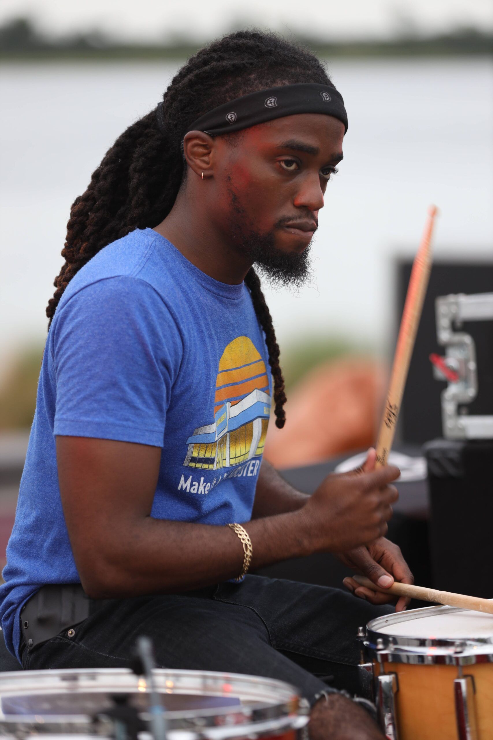 Drummer in a blue shirt playing at an outdoor performance near water