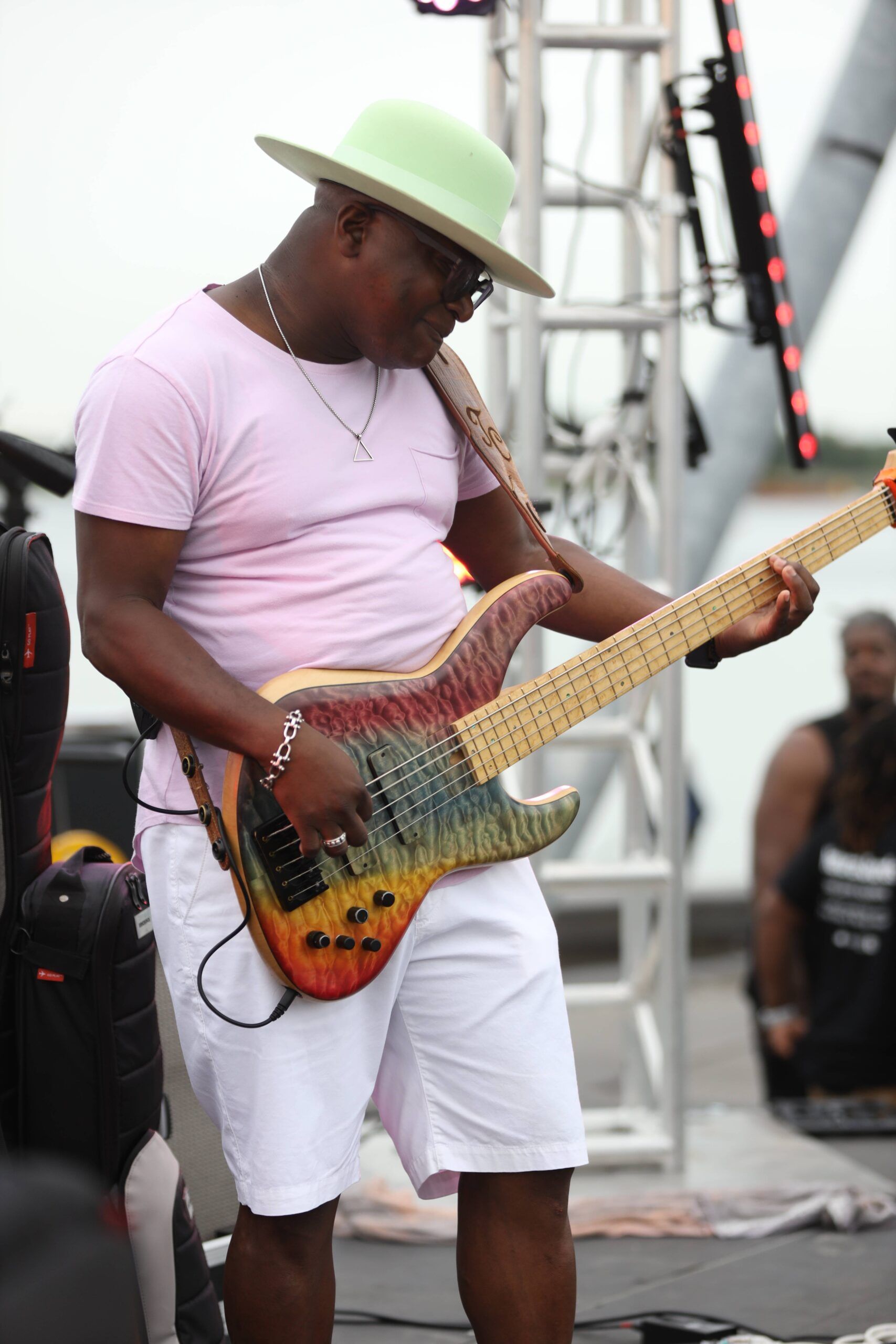 Musician playing an electric bass guitar on an outdoor stage, wearing a white hat and white clothes.