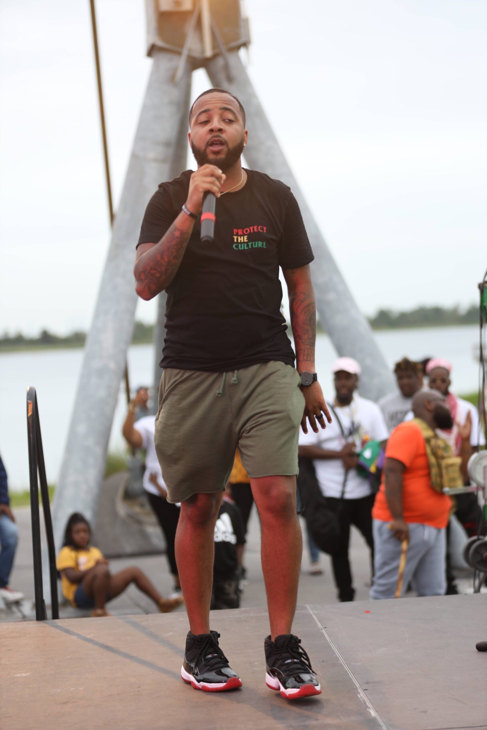 Man speaking into a microphone on an outdoor stage with people gathered behind him.