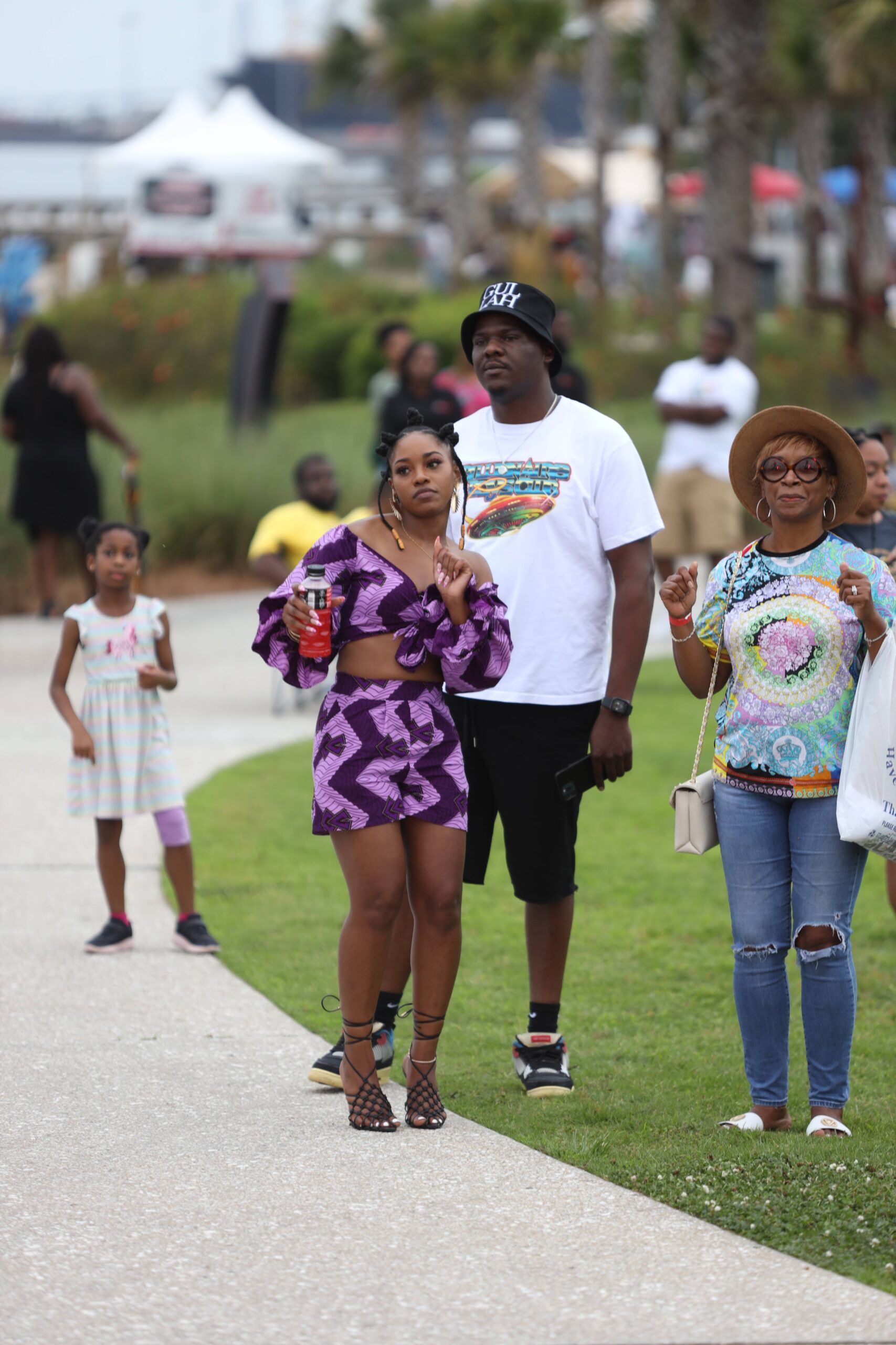 Three people walking on a park path; one gestures while others smile, with palm trees and grass in the background.