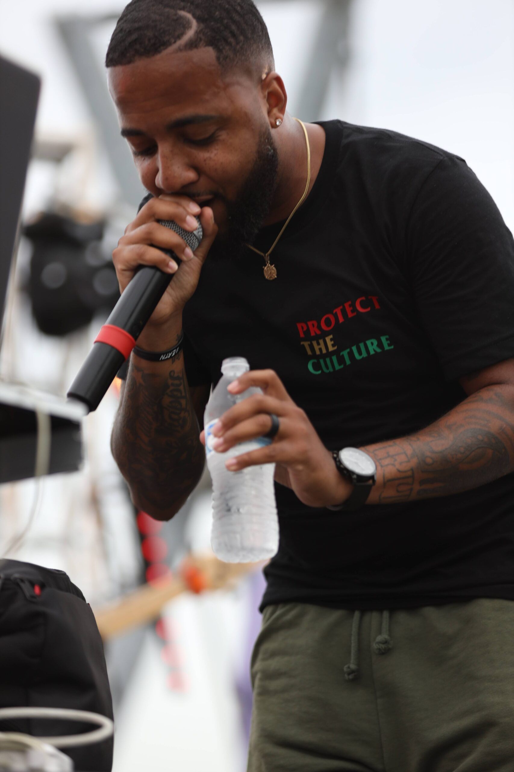 Man singing into a microphone while holding a water bottle, wearing a black shirt and olive pants on stage