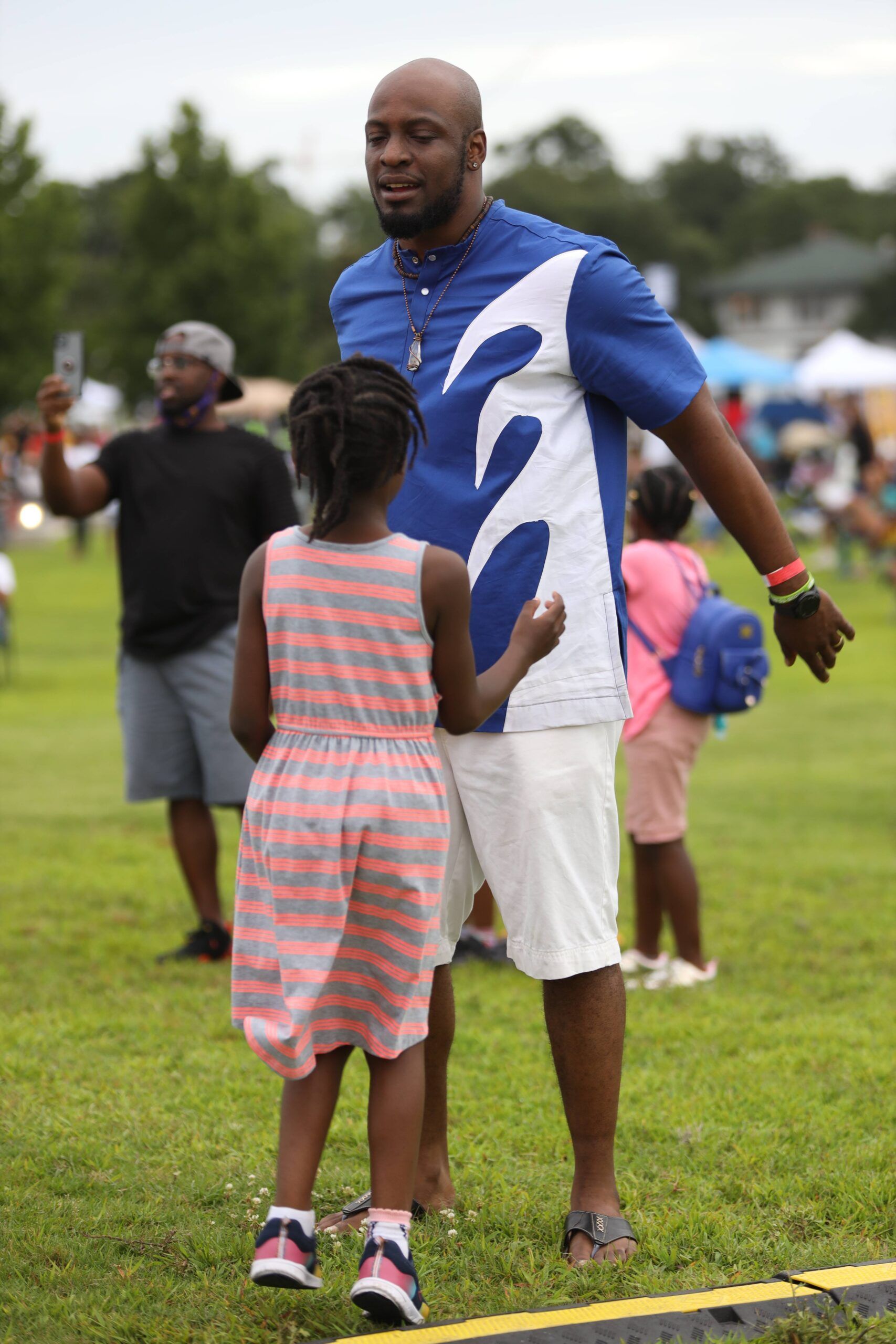 Man in blue shirt standing on a grassy field with a child in a striped dress, outdoors at a park event
