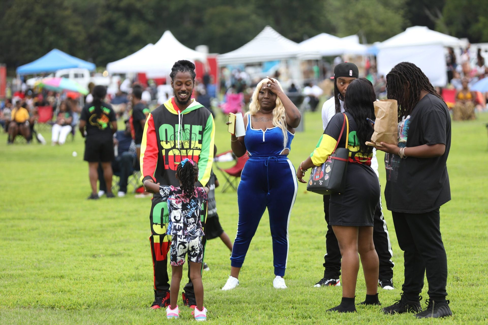 People standing on a grassy field at an outdoor event with tents and crowds in the background.
