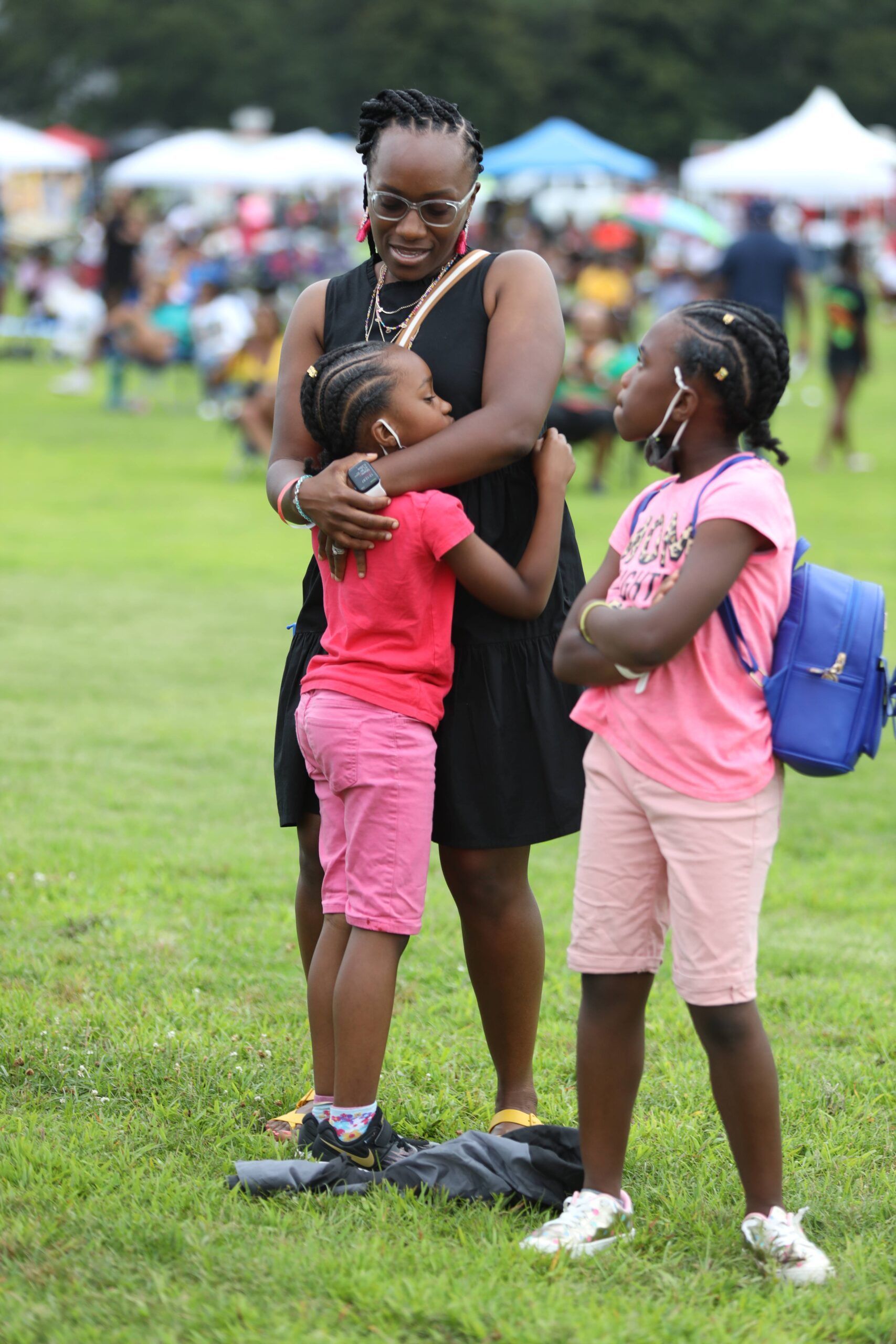 Two children hugging a smiling adult in a grassy park, with tents blurred in the background.