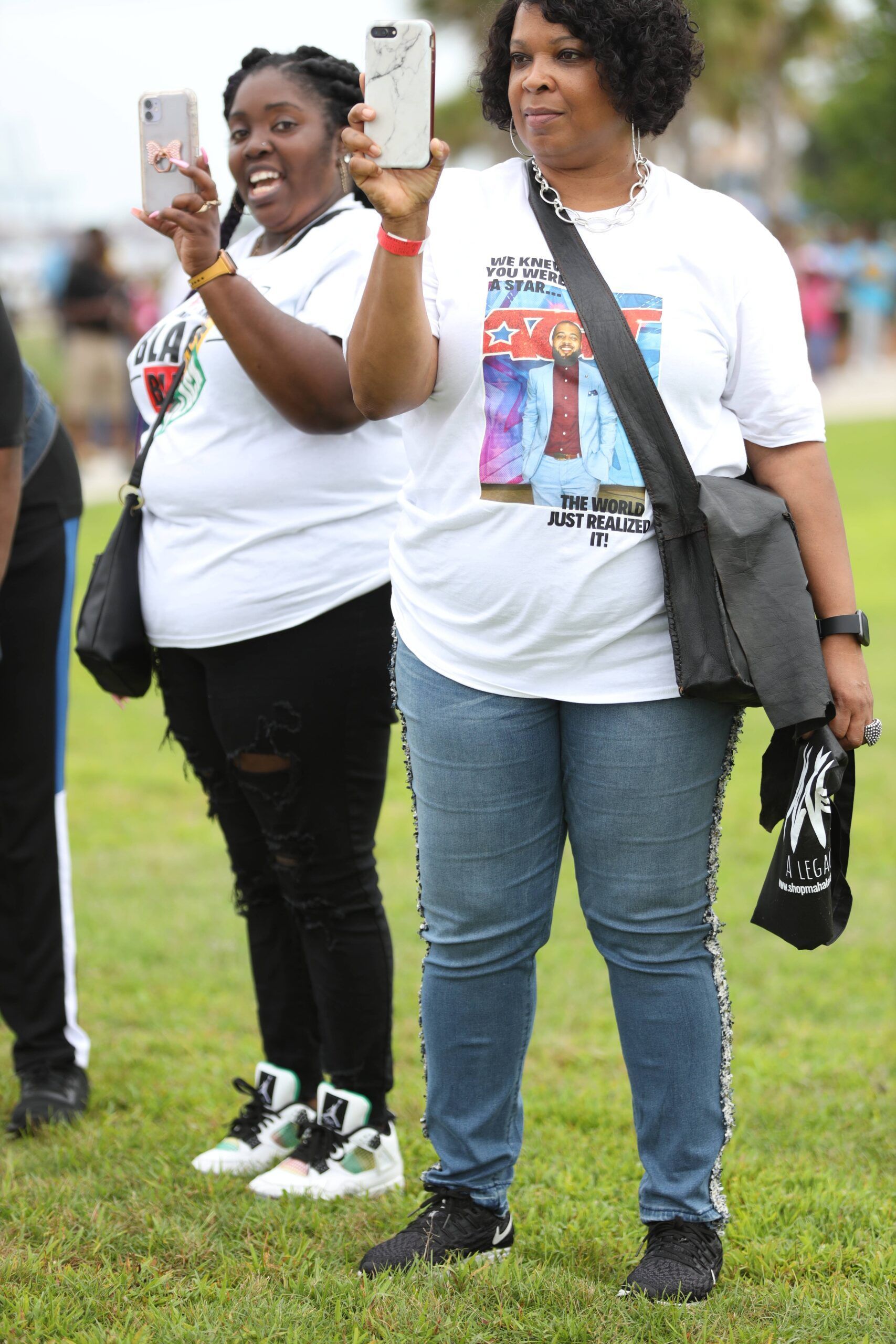 Two people in white T-shirts pose on a grassy field, one taking a selfie with a phone.