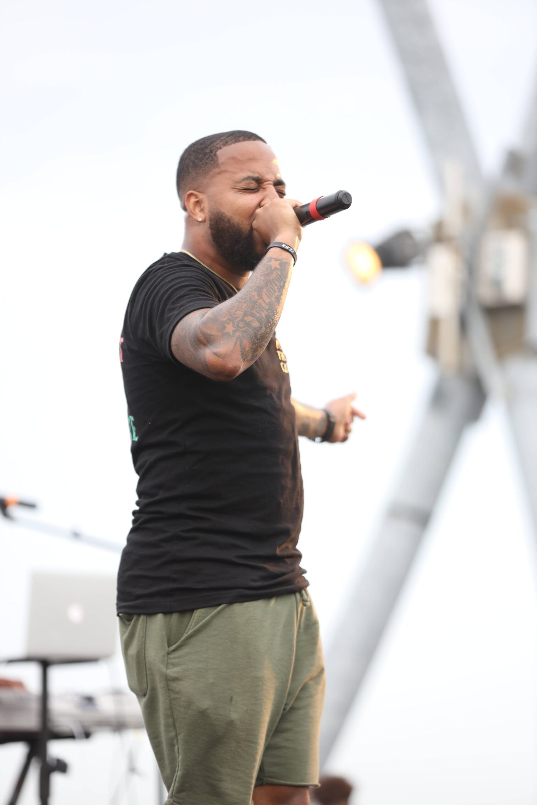 Singer holding a microphone onstage, wearing a black T-shirt and olive shorts.