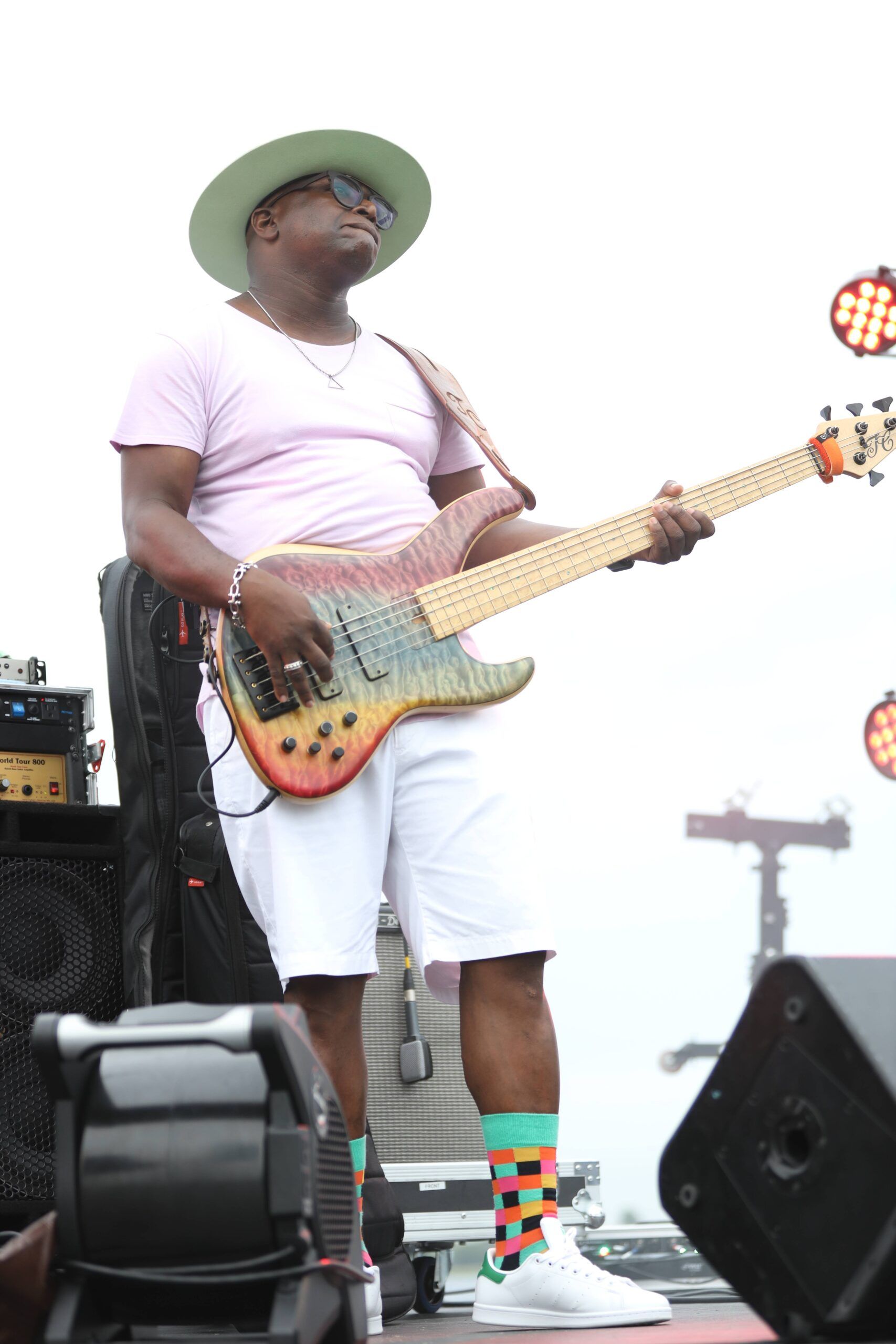 Musician in pink shirt and white shorts playing bass guitar onstage, wearing a green hat and colorful socks