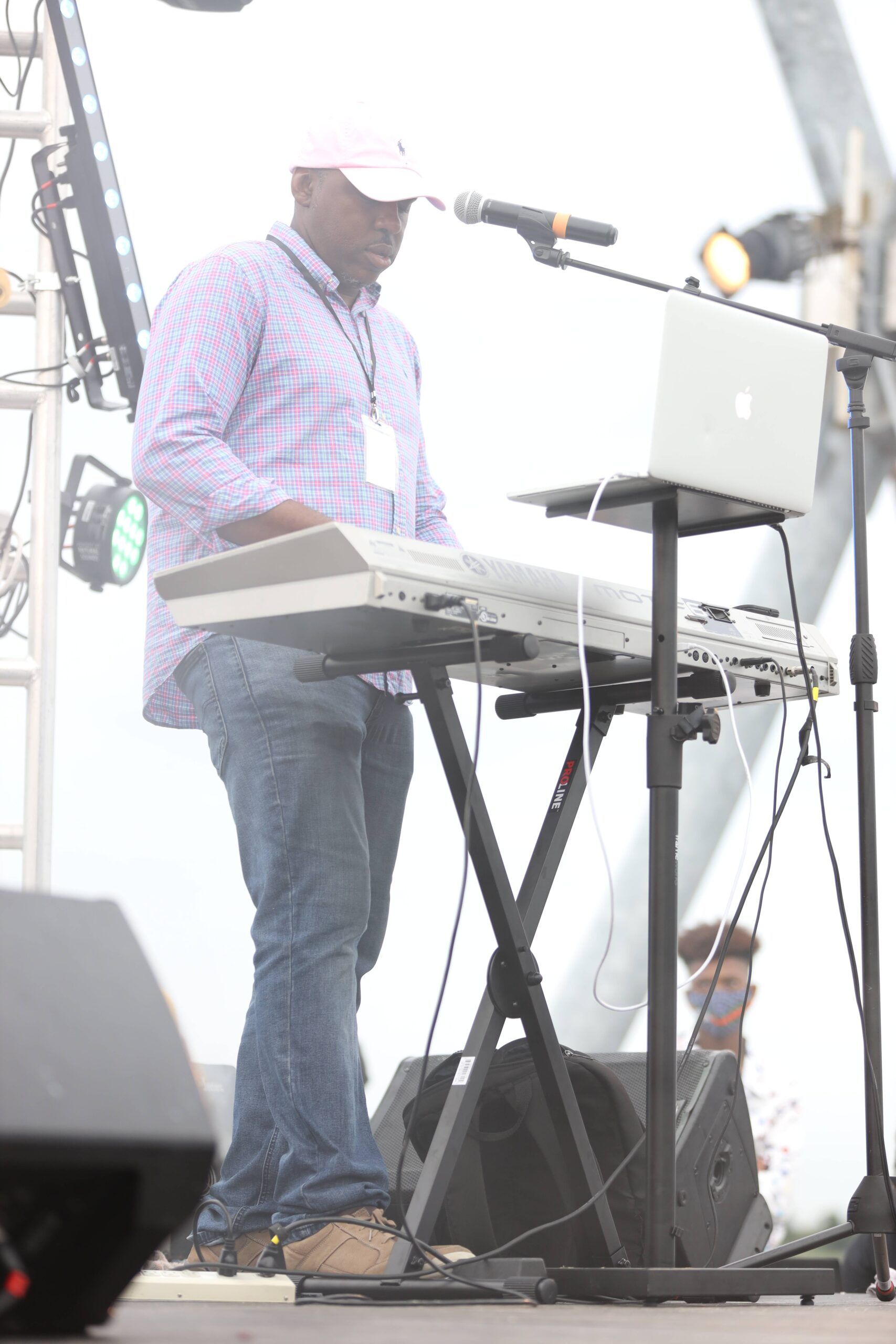 Musician playing keyboard on an outdoor stage beside a laptop and microphone stand