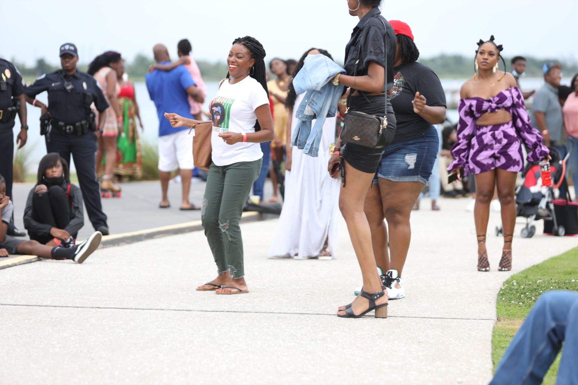 Crowd standing and walking on a waterfront promenade, with people in casual clothes and a clear sky.