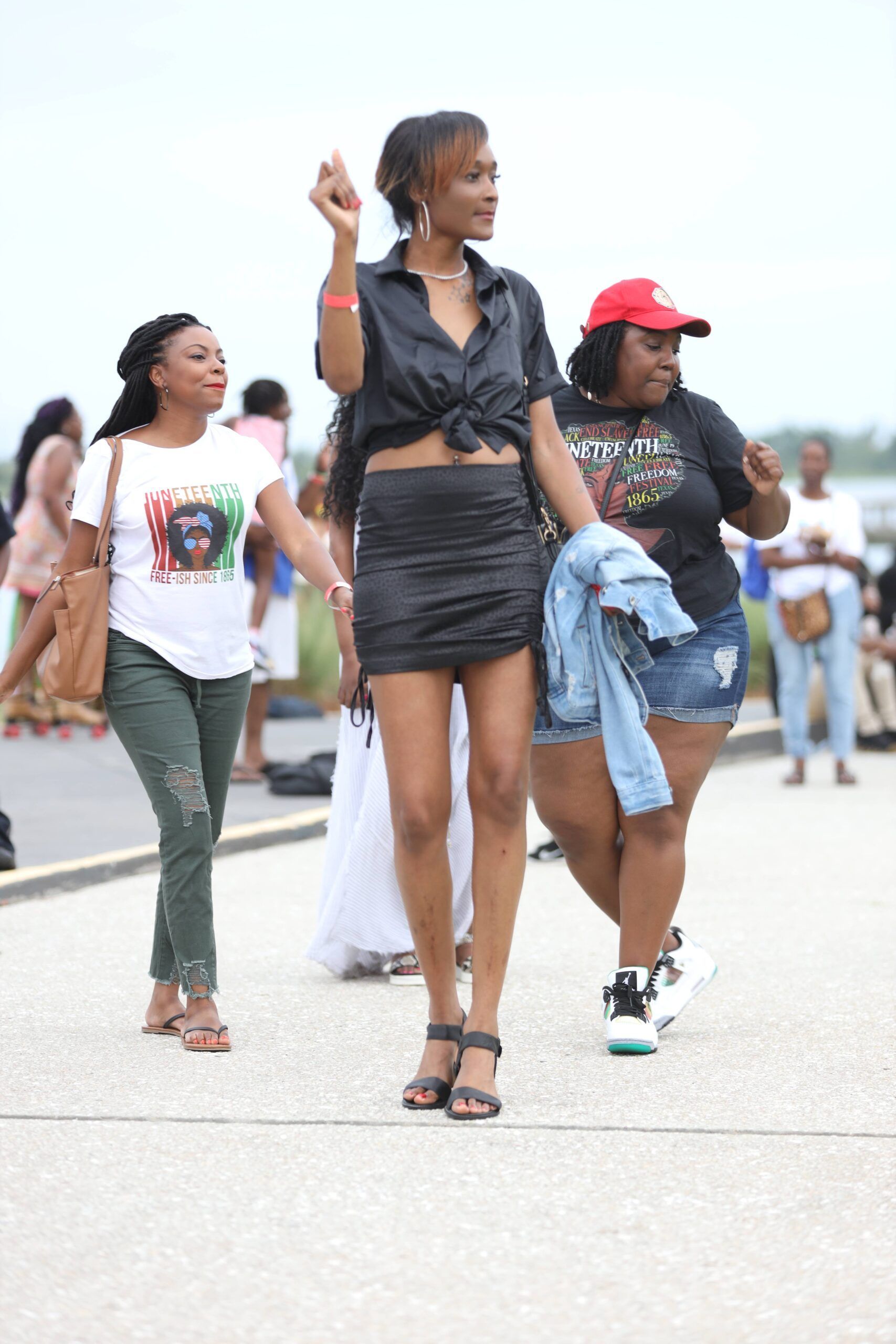 Three women walking outdoors on a paved street, one in black holding a cup aloft, others in casual clothes.
