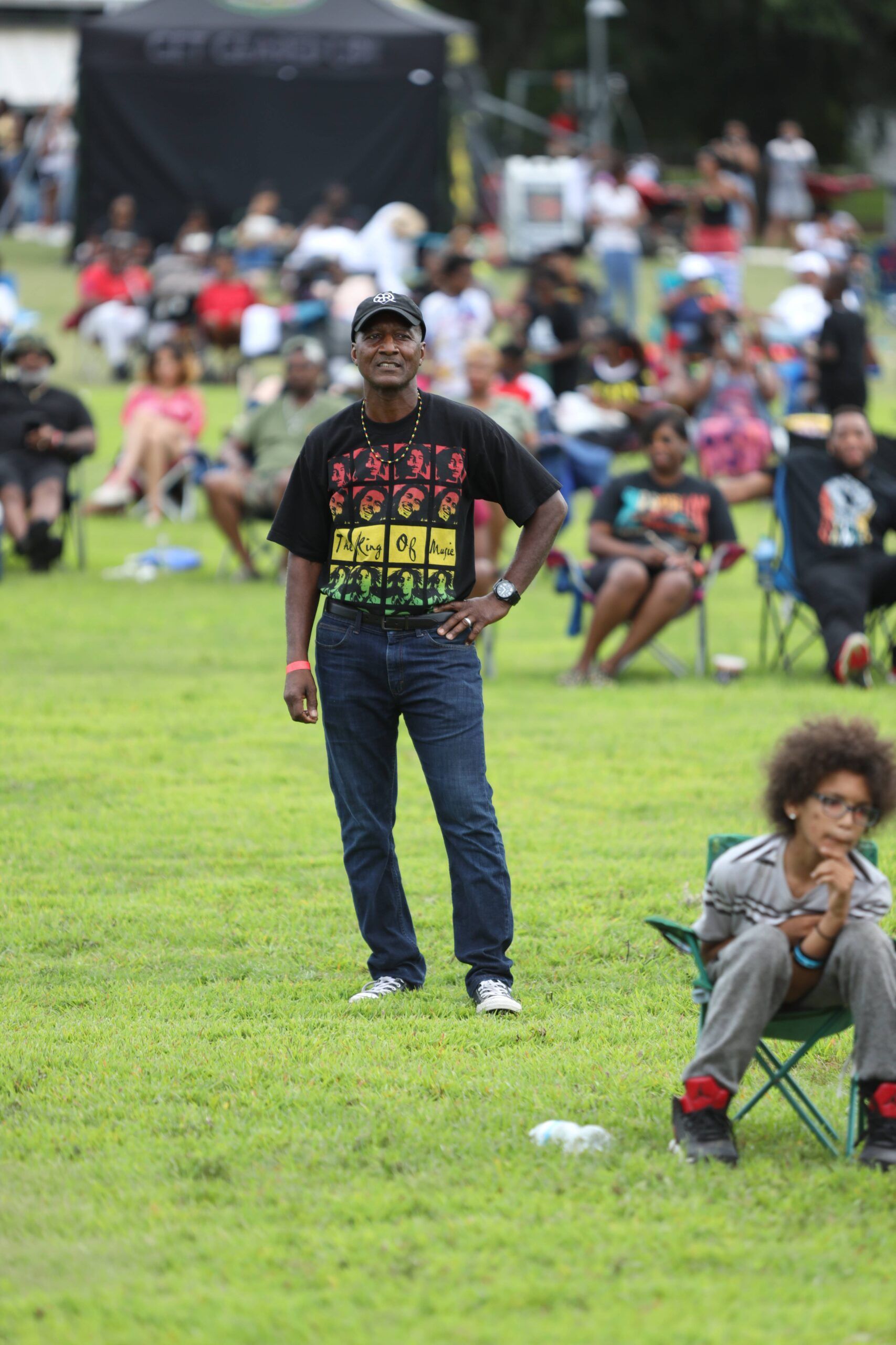 Man standing on a grassy field at an outdoor event, with a seated child in the foreground and a crowd behind.
