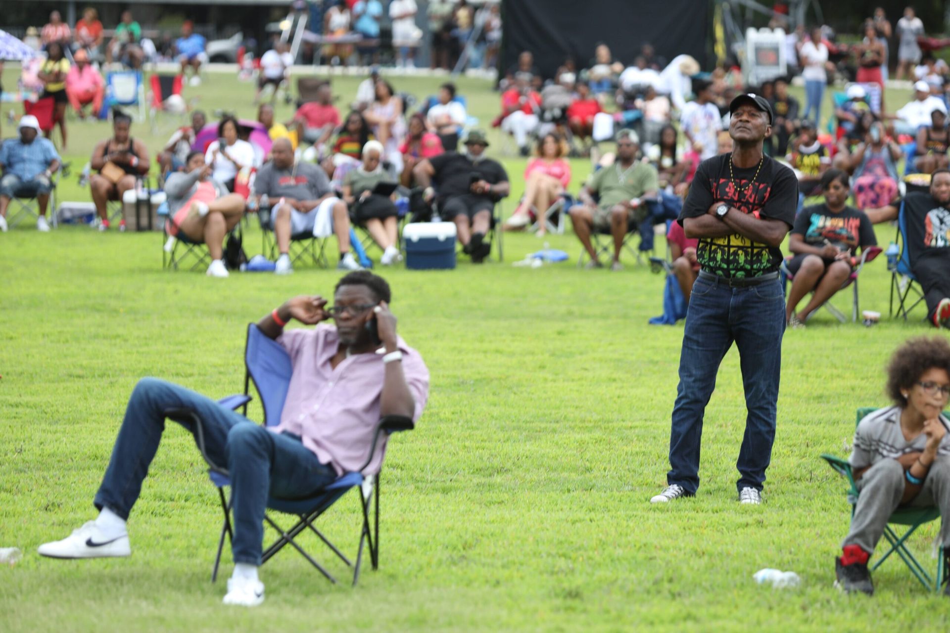 People sitting on folding chairs on a grassy field at an outdoor event, with a crowd in the background.
