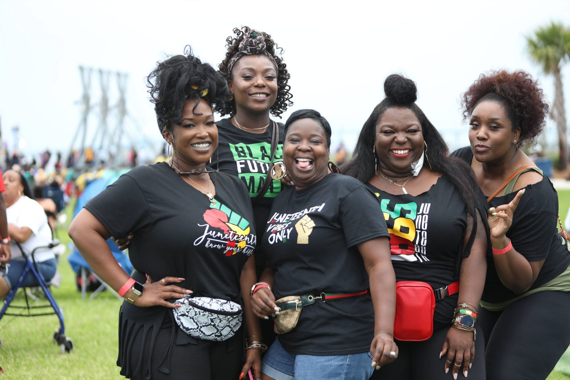 Five smiling friends in black T-shirts posing together at an outdoor event on a grassy field.