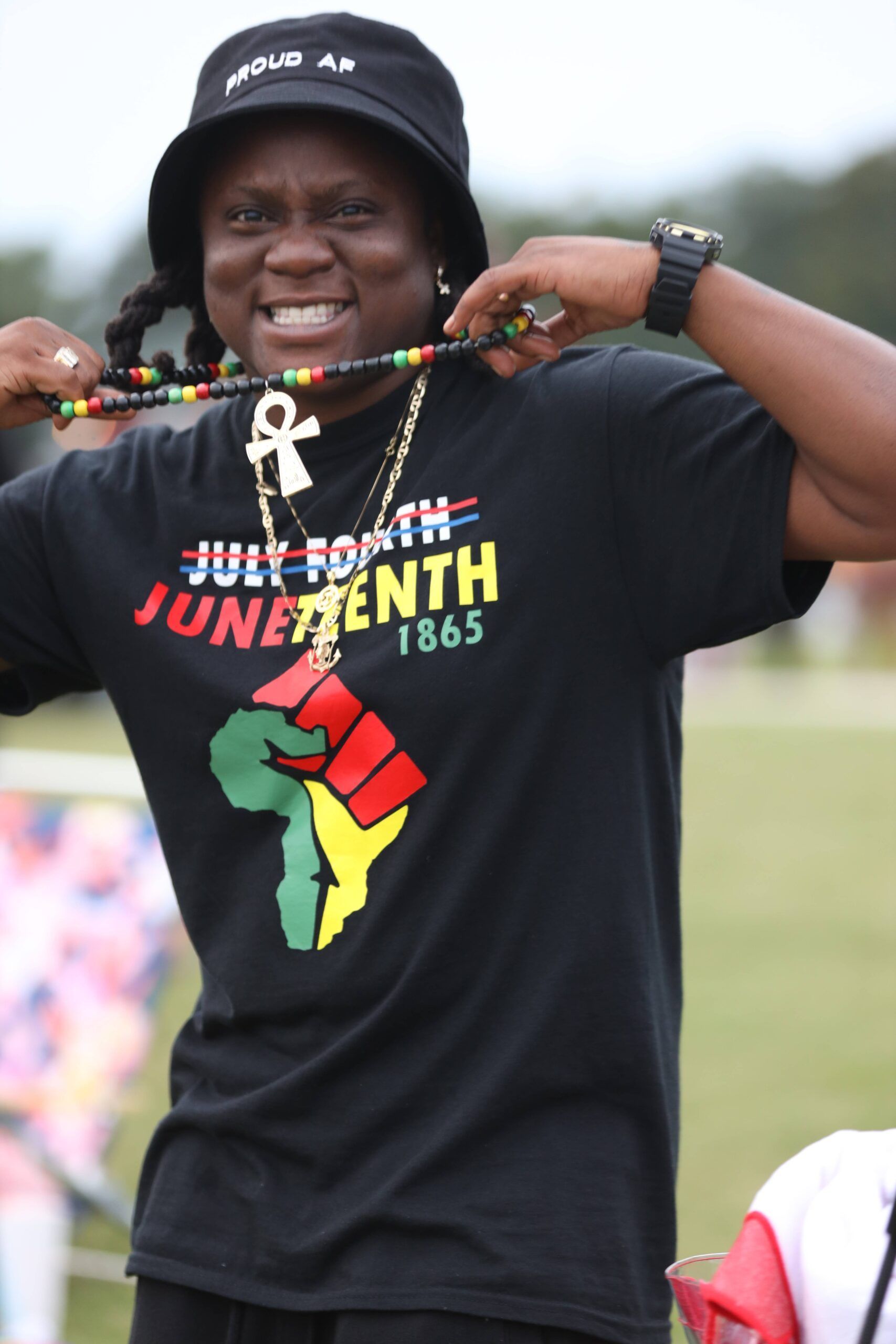 Person in black Juneteenth T-shirt smiling and holding beaded necklaces outdoors
